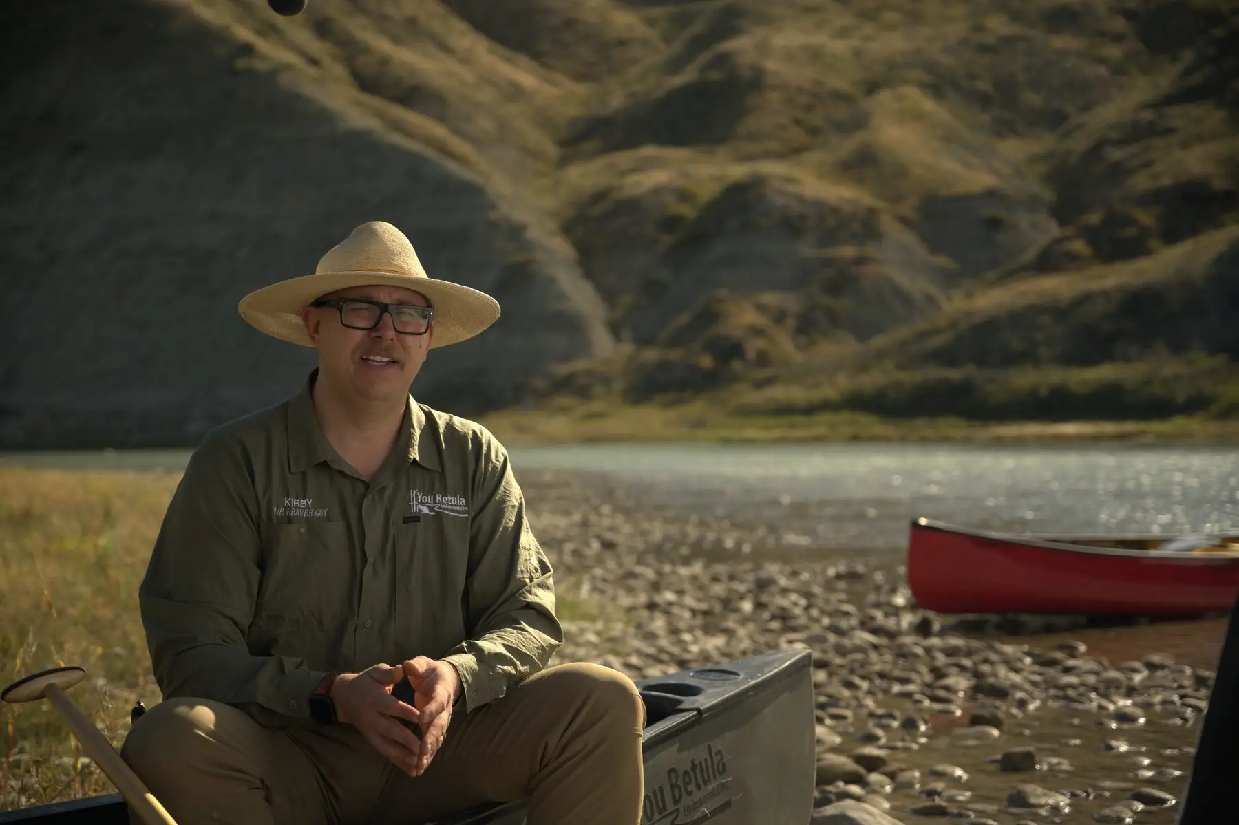 Kirby England seated beside a red canoe on a gravel bar